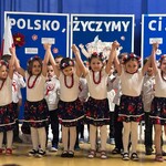 Odświętnie ubrane dzieci stoją z uniesionymi rękami, a dziewczynki mają na sobie korale oraz wianki na włosach.