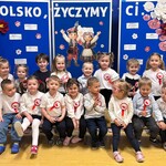 Dzieci odświętnie ubrane siedzą na tle tablicy z napisem „Polsko, życzymy Ci…”.